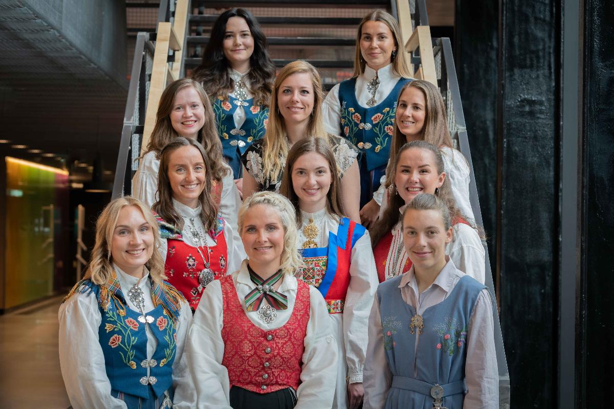 Gruppebilde av 11 jenter med ulike bunader som står oppstilt i en trapp 