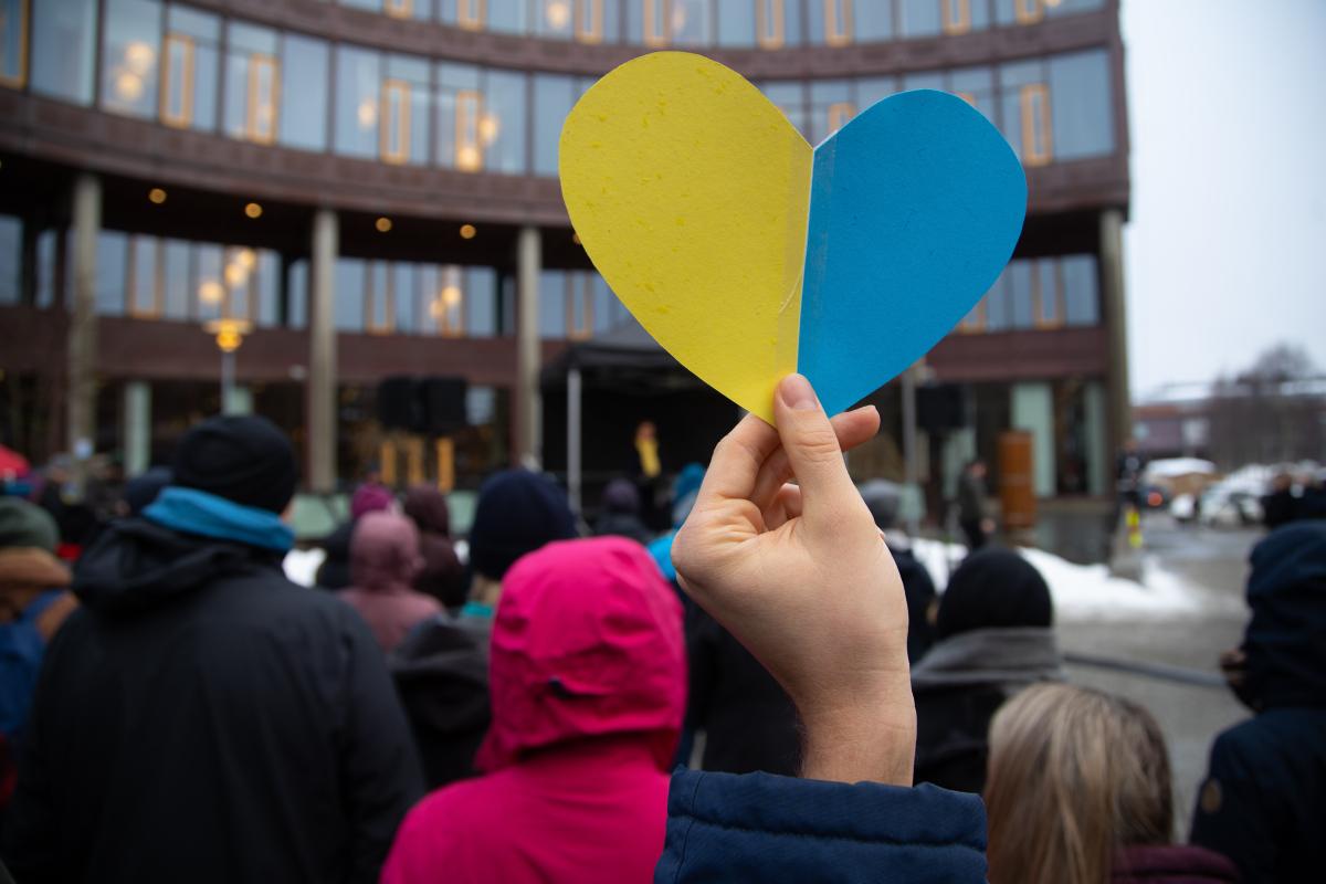 A paper heart in yellow and blue held up by a hand in a crowd outside. 