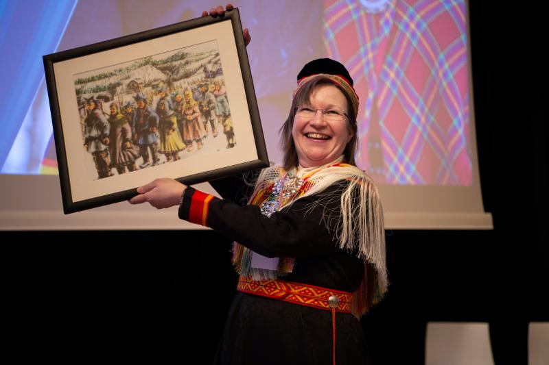 Ei glad kvinne på ei scene held opp eit bilete/kunstverk. Ho er kledd i samsik kofte.