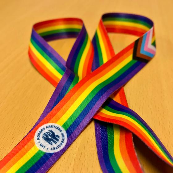 A rainbow-coloured lanyard draped on a table, with the UiT logo in the foreground, and the black, brown, blue, pink and white progress stripes in a V-pattern prominent on the rainbow stripes.