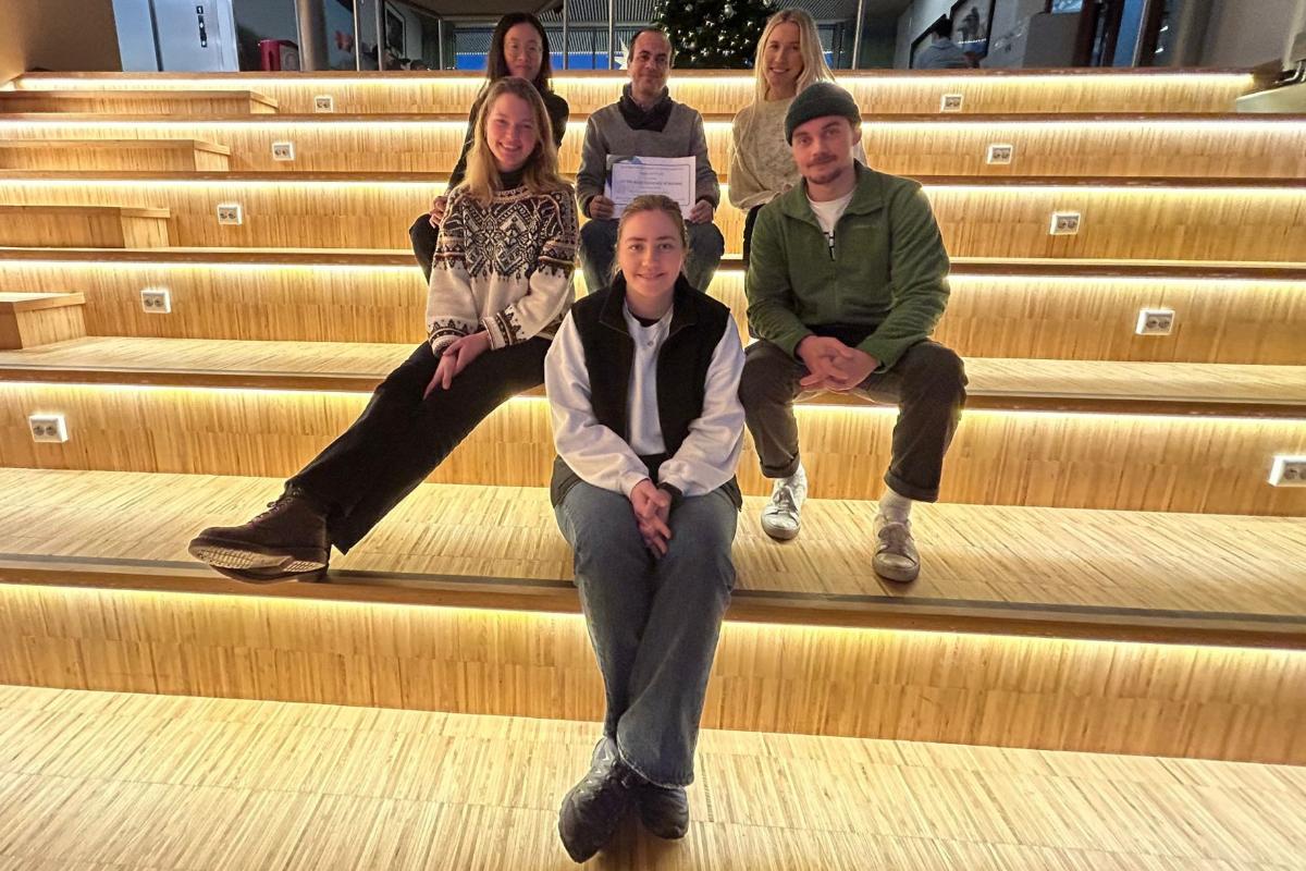 seks menneske sit smilande i ei opplyst, innand&oslash;rs trapp. Mannen i midten held eit diplom. juletre i bakgrunnen.