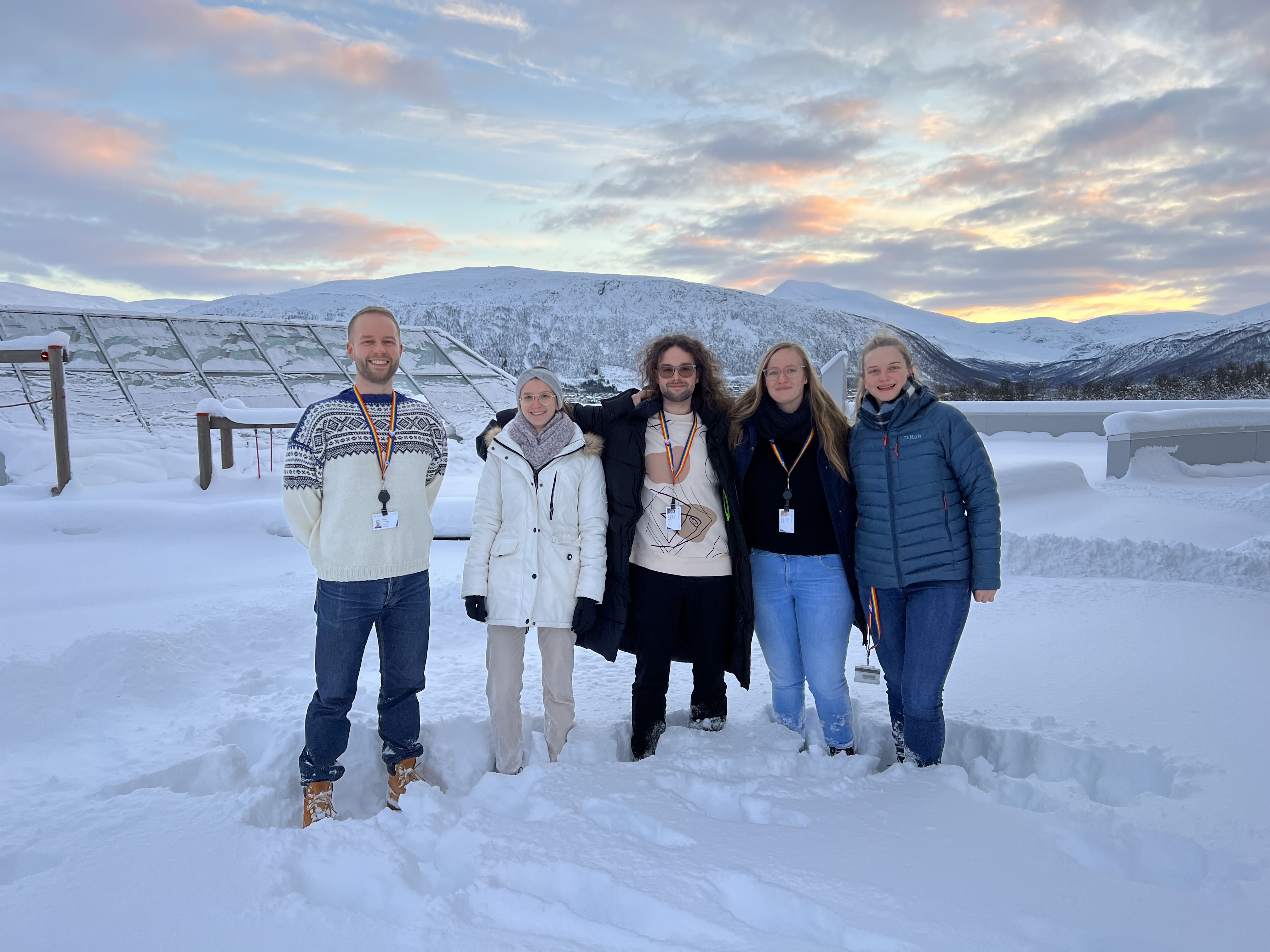 The Haugland research group in 2023. From left to right: Marius, Lea, Mateusz, Floriane, Anna. They are smiling, standing in the snow, with a view of the sea, snow-clad mountains and colourful, polar night skies.