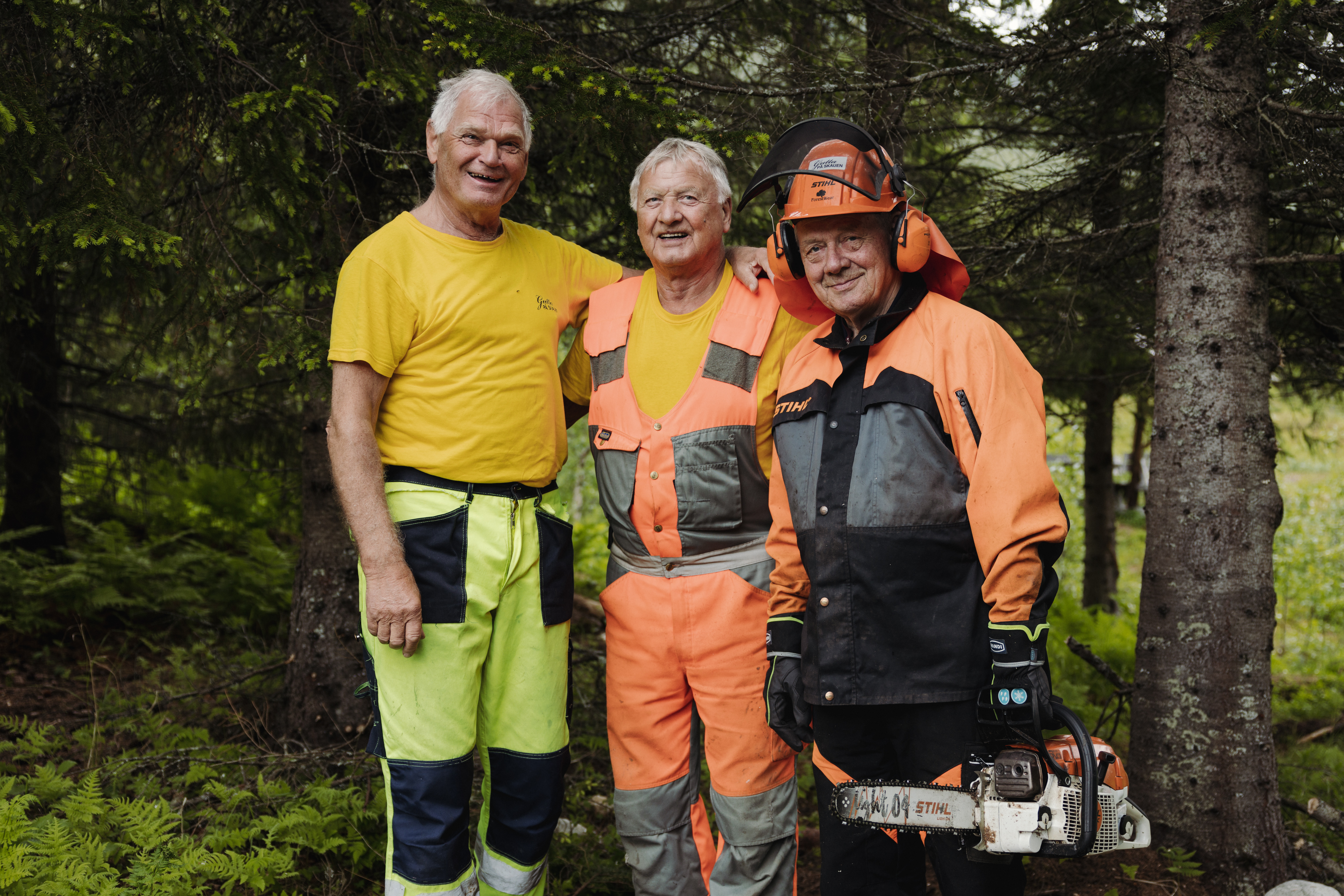 tre eldre menn som jobber i skogen med arbeidsklær og motorsag