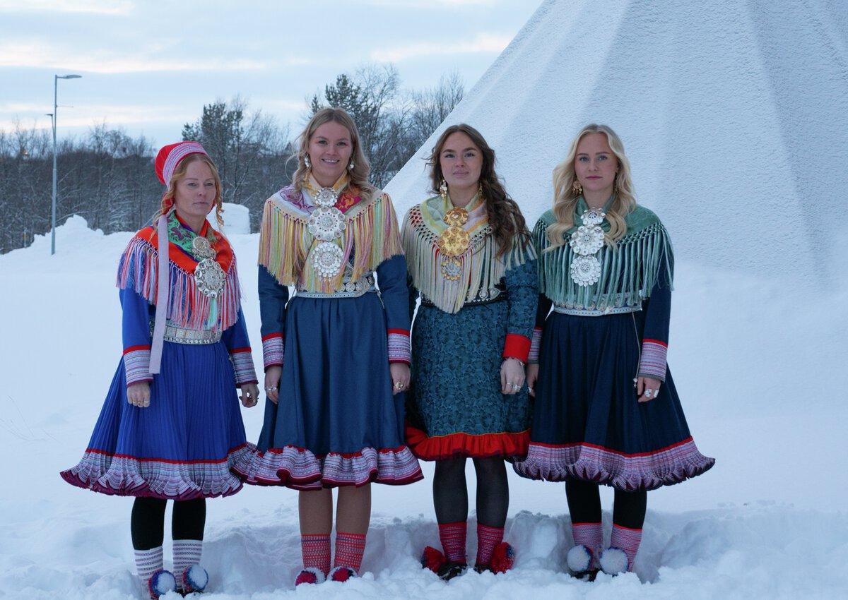 Fire unge kvinner i ulike samiske kofter poserer foran en lavvo. V&aelig;ret er kaldt og klart.