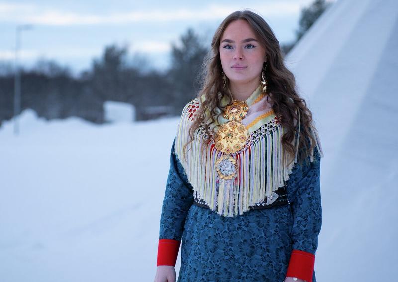 En ung kvinne i samisk kofter poserer foran en lavvo. V&aelig;ret er kaldt og klart.
