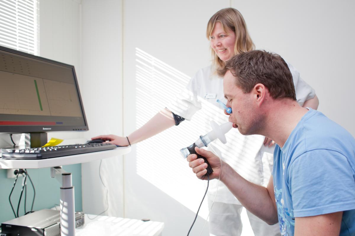 en mann bl&aring;ser i et medisinsk apparat. en kvinne i sykehuskl&aelig;r st&aring;r i bakgrunnen og ser p&aring;