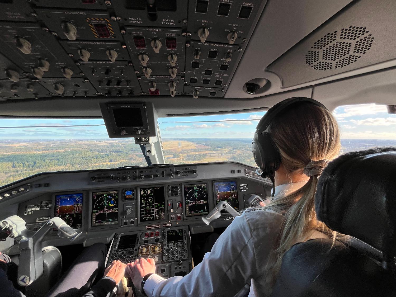 En kvinnelig pilot sett bakfra mens hun styrer et fly, med et vidstrakt landskap synlig gjennom cockpitvinduene.