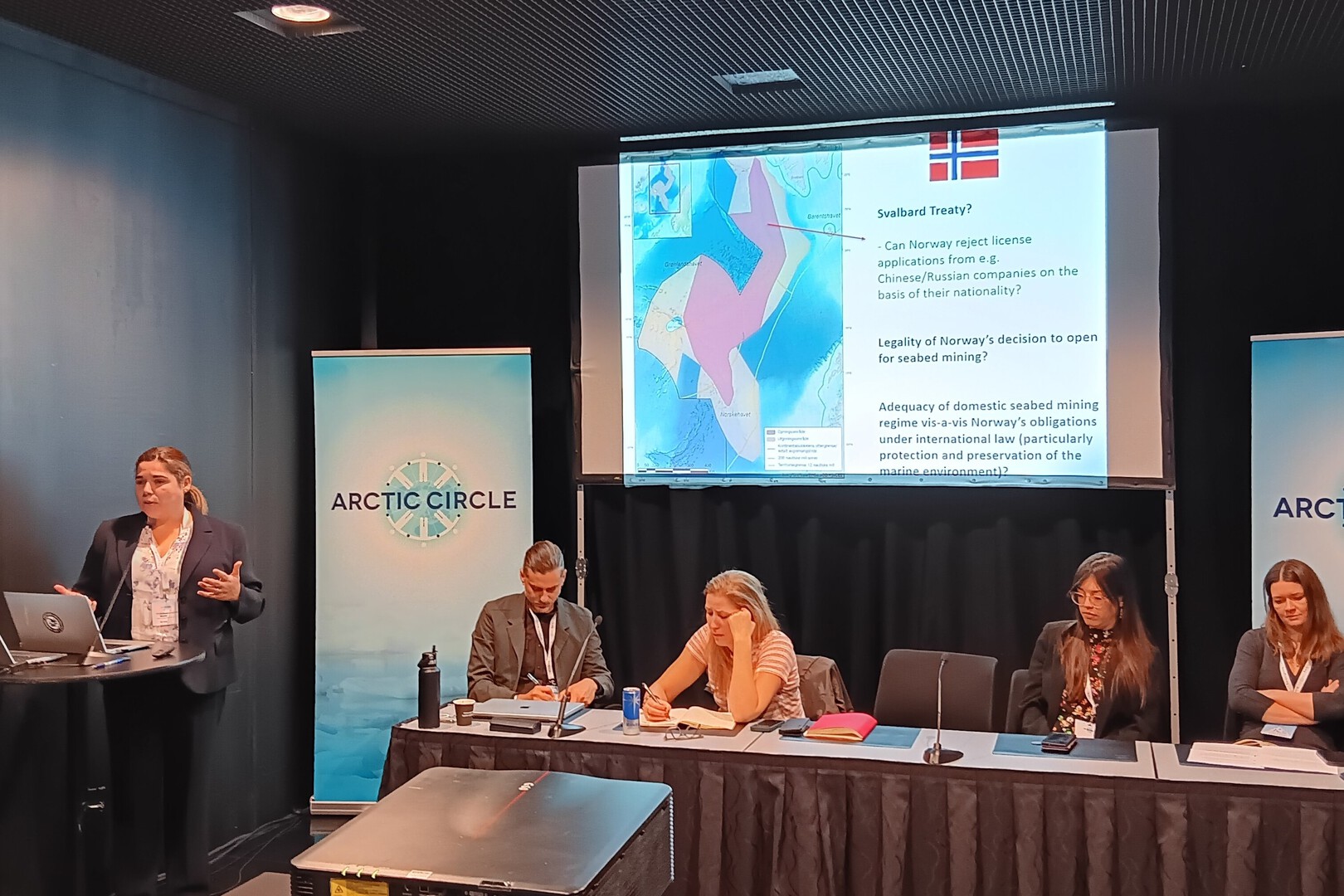 One speaker, with several others gathered around a table, below a projector screen.