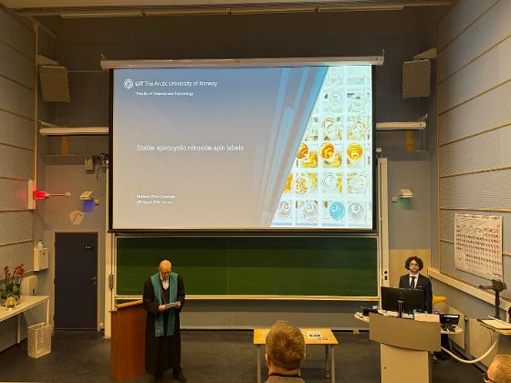 Mateusz standing at the lectern ready to begin the presentation of his PhD work.