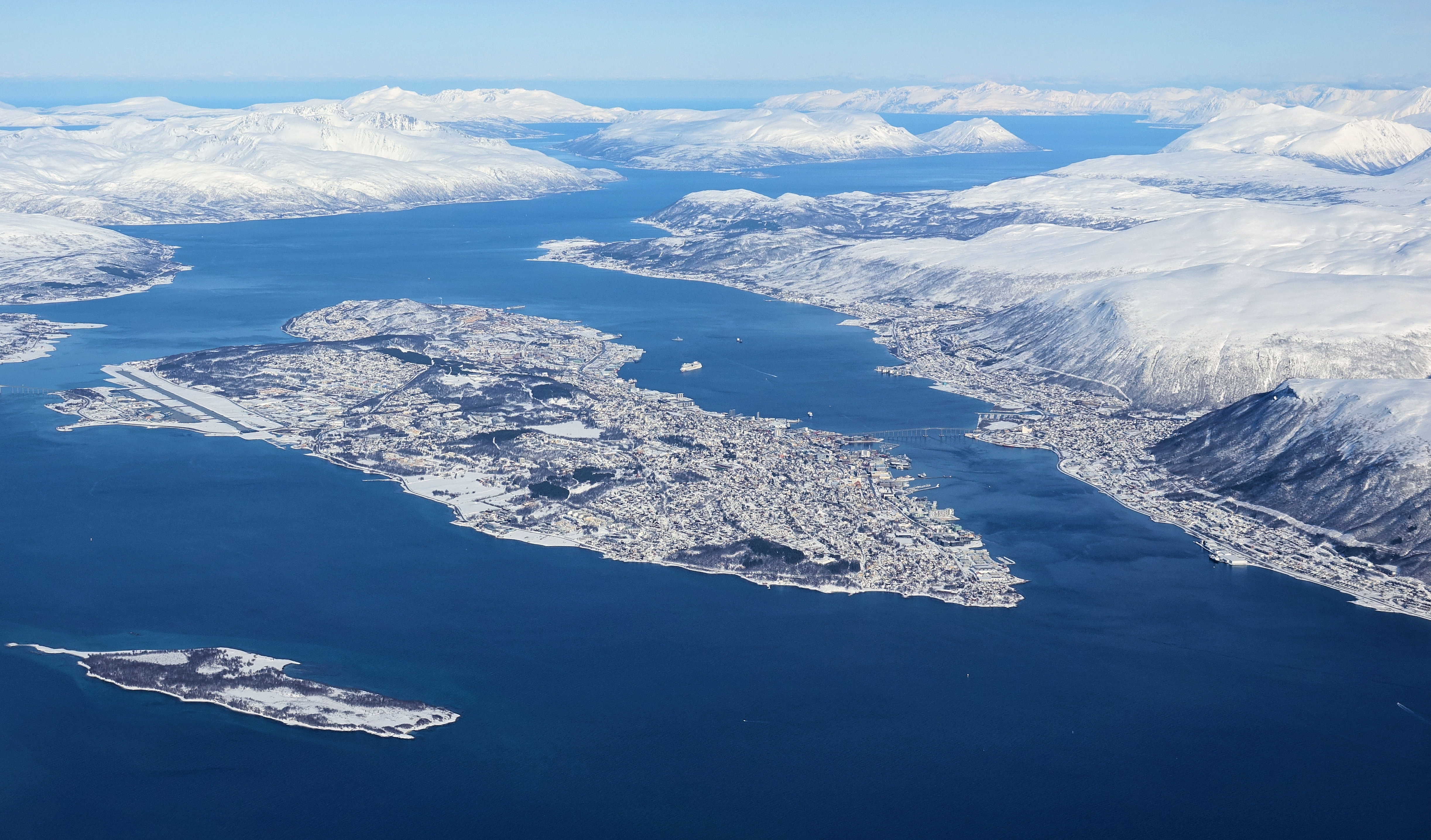 Tromsøya island with the smaller Store Grindøya island in the lower left foreground. Most of the city of Tromsø is located on Tromsøya, and the city continues across the bridges on the the east (Tromsdalen on the mainland) and west (Kvaløya island) side of the island. Picture by Bjørn Christian Tørrissen, licensed under the Creative Commons Attribution-Share Alike 4.0 International license.