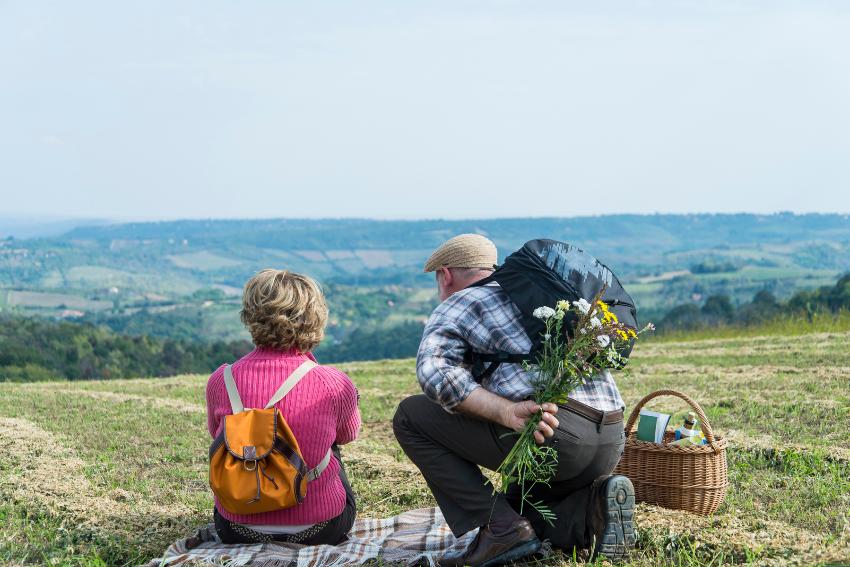 Et eldre par sitter på et teppe på en slette med ryggen til og ser ut over et grønt landskap. Mannen har en bukett med blomster bak ryggen.