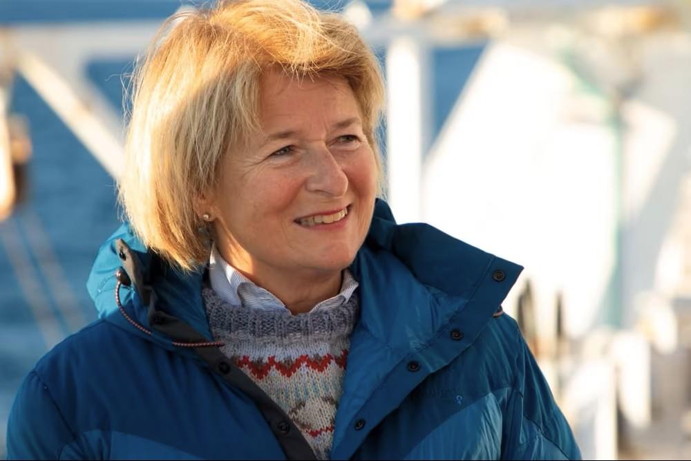 Dame med blondt hår som smiler og har på dunjakke. Det er ute. 