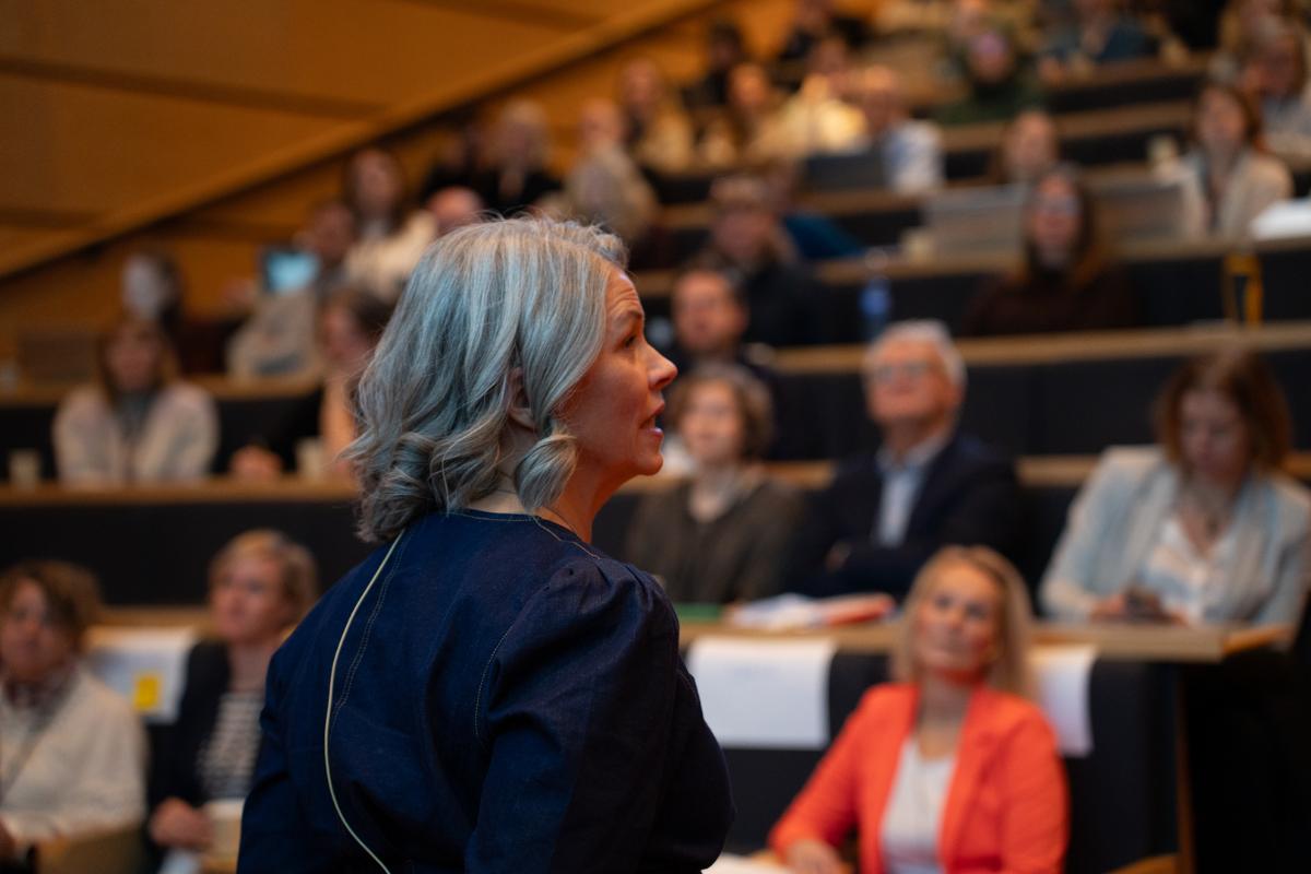 Kvinne taler til forsamling i forgrunnen. 