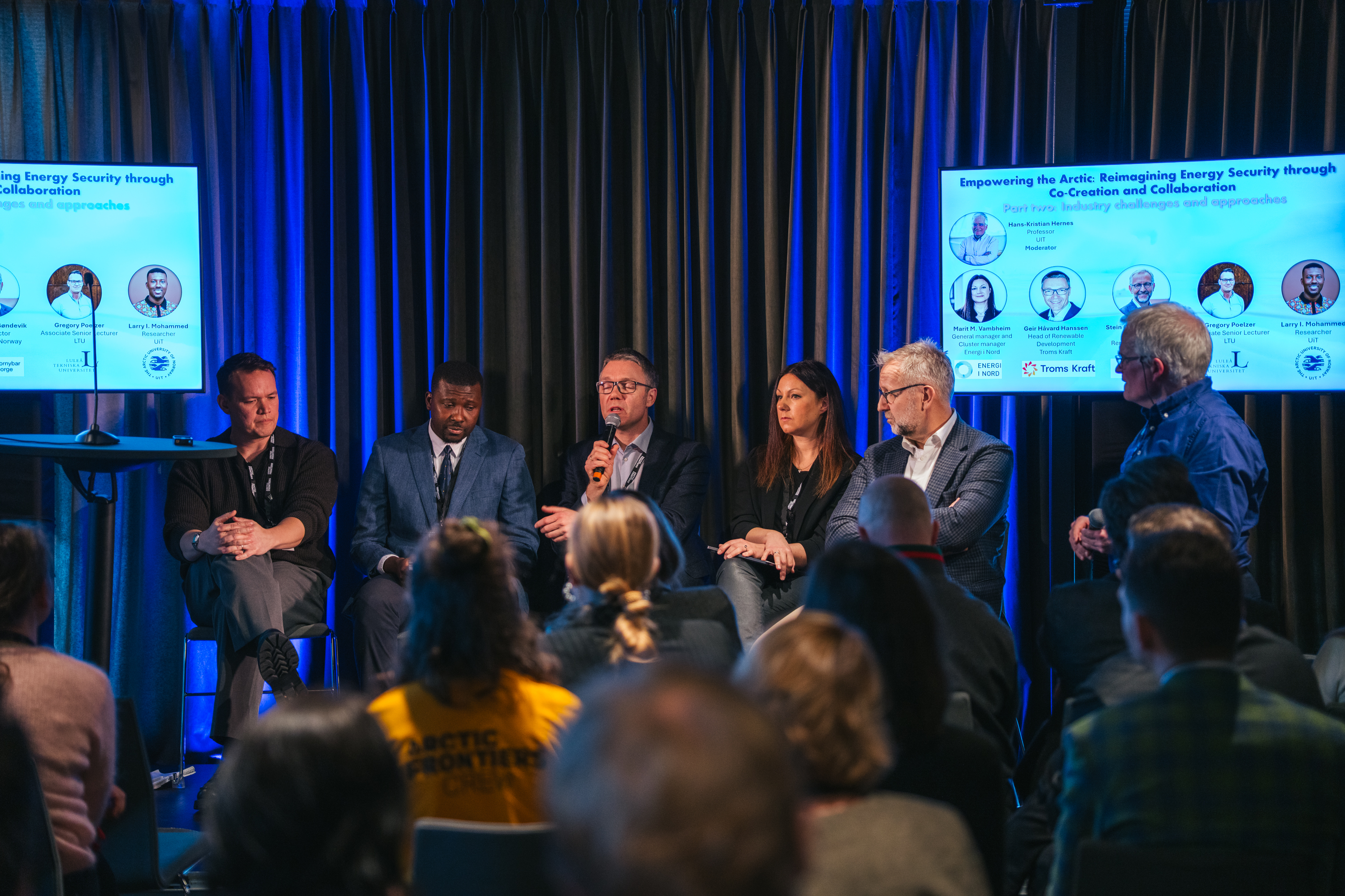 Folk som sitter p&aring; en scene og diskuterer, mellom to skjermer.
