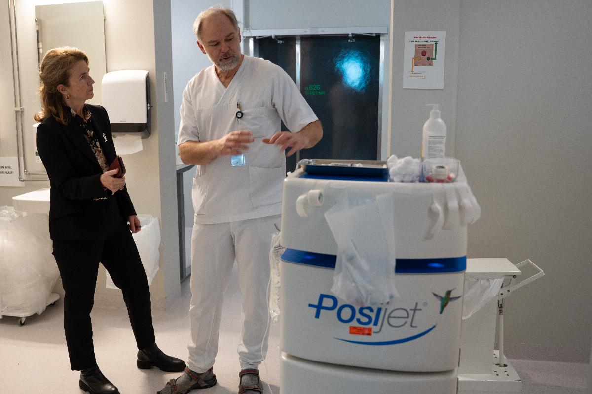 en mann i sykehus-antrekk forklarer noe for en kvinne foran en stor medisinsk maskin