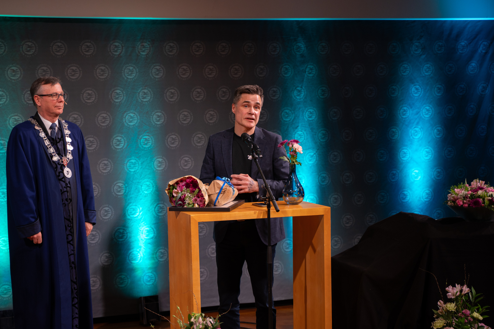 Robert Jenssen-03662.jpg En mann mottar diplom, blomster og gave i staselig og feststemt universitetsauditorium.