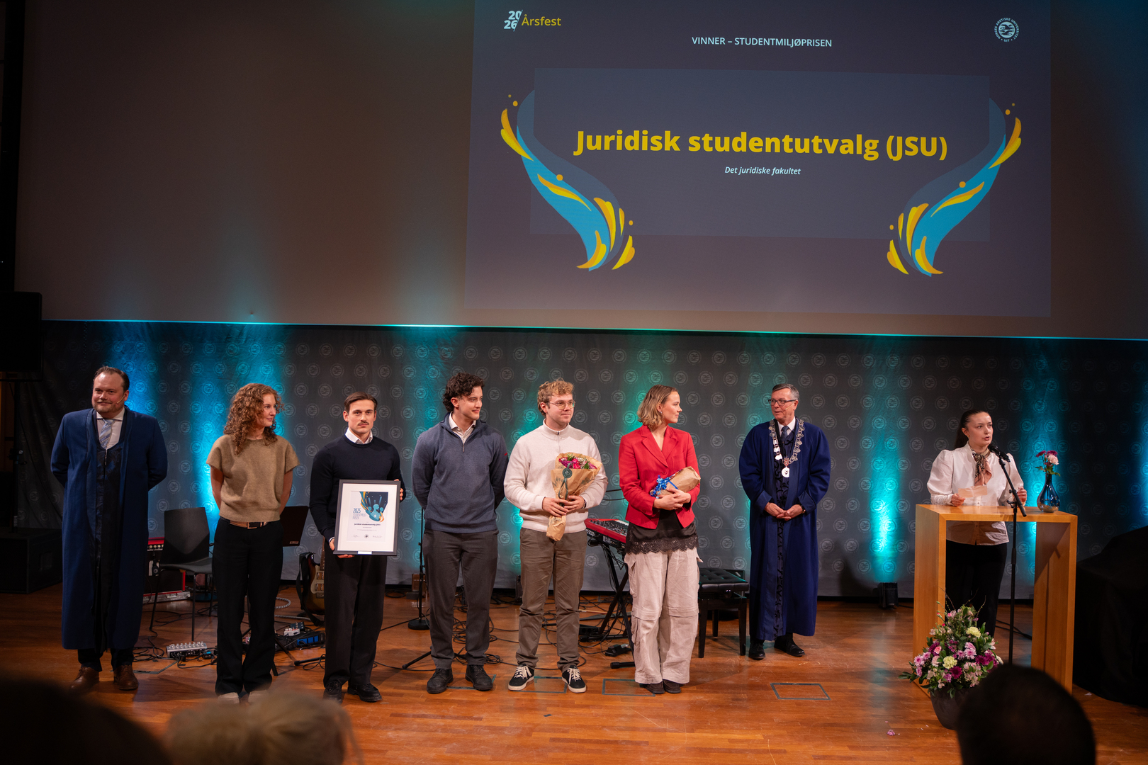 Studentmiljøprisen-03719.jpg En gruppe entusiastiske unge studenter og ansatte mottar diplom, blomster og gave i staselig og feststemt universitetsauditorium.