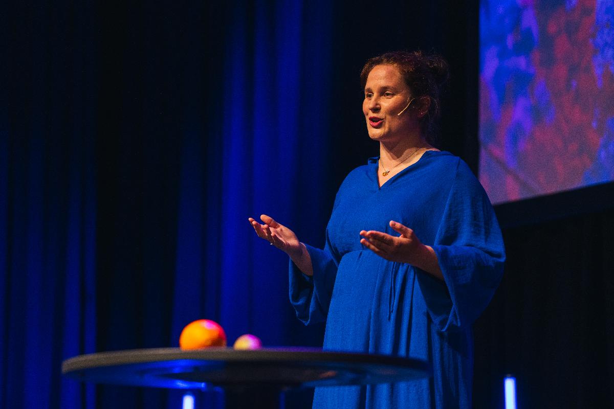 KVinne i bl&aring;tt st&aring;r p&aring; en scene i bl&aring;tt lys, med en appelsin i h&aring;nden.