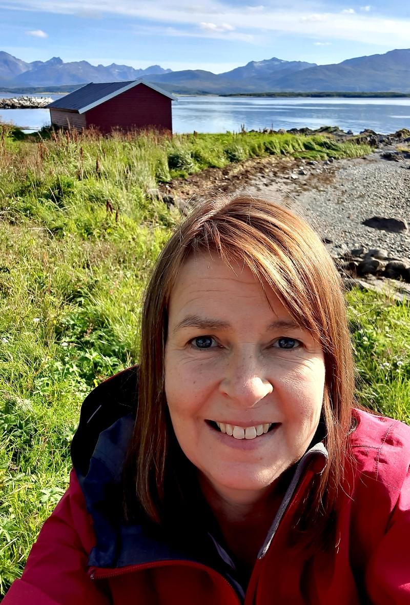 Tiina.jpg dame som står i fjæra med hav og naust i bakgrunnen