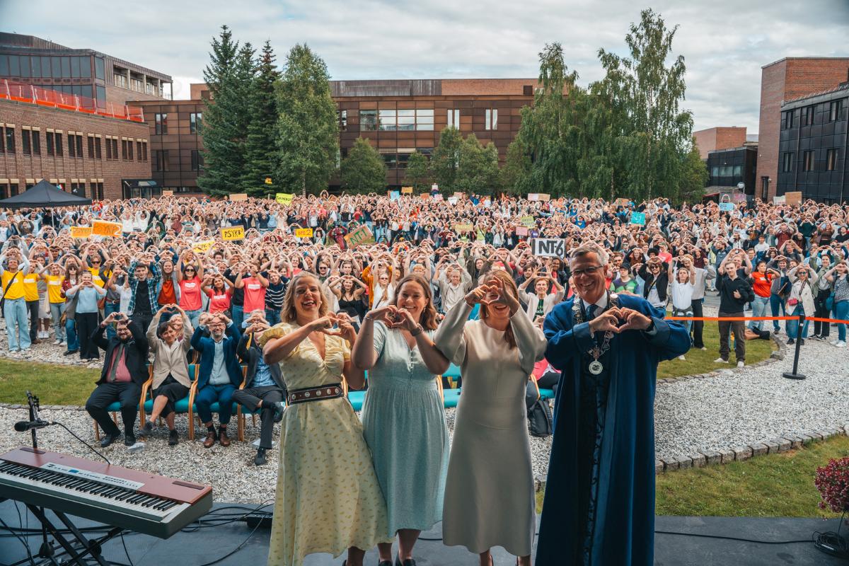 Et knippe rektorer og politikere viser hjertetegn med hendene p&aring; en scene med masse glade, forventningsfulle studenter i bakgrunnen.  