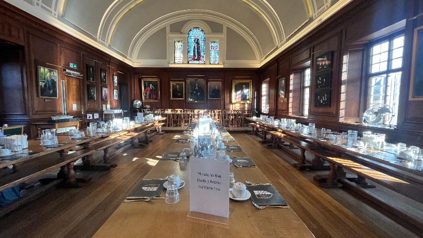 A wood-panelled Oxbridge-style dining hall set for a formal meal, with long refectory tables laid with menus, glasses and coffee cups. Oil portraits in gilt frames line the panelled walls beneath a barrel-vaulted ceiling and a stained-glass window. On the nearest table, a printed sign reads 'Music in the (sub-) Arctic conference'.